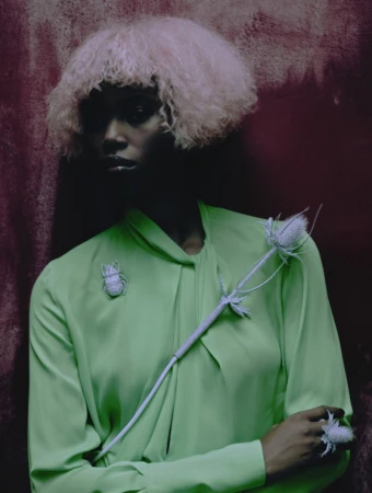 Dark-skinned woman poses against a red wall wearing a green blouse, insect brooch, white wig, and flower ring in a high-fashion setting.