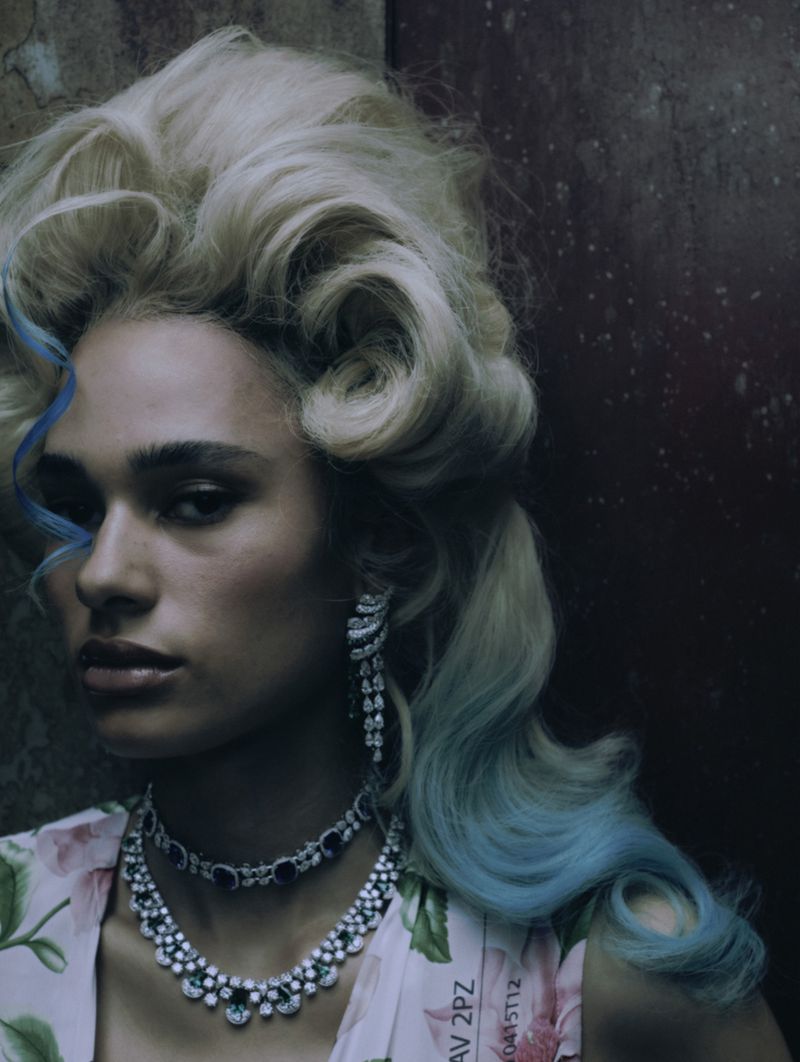 Close-up of a woman with blond hair styled in an updo and turquoise highlights, wearing jewelry and a floral print top.