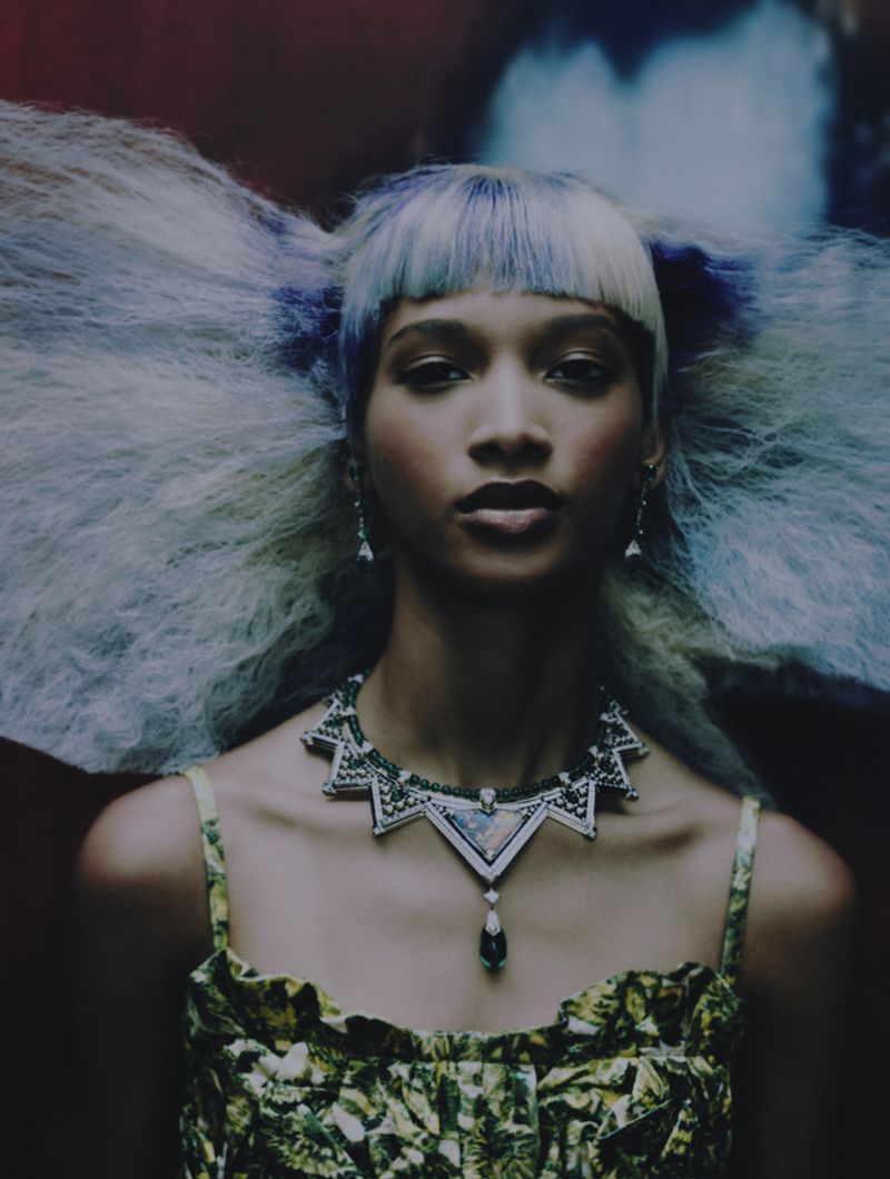 Portrait of a woman with blue and white hair, wearing a patterned green top and an elaborate necklace, against a dark background.