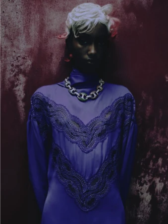 Model with sculpted white hair wearing a high-neck purple lace dress and chunky chain necklace against a textured crimson backdrop.