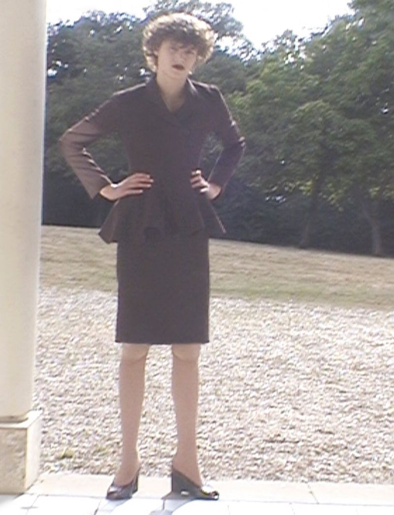 Adult female in a brown skirt suit standing on a stone patio with trees in the background on a sunny day.