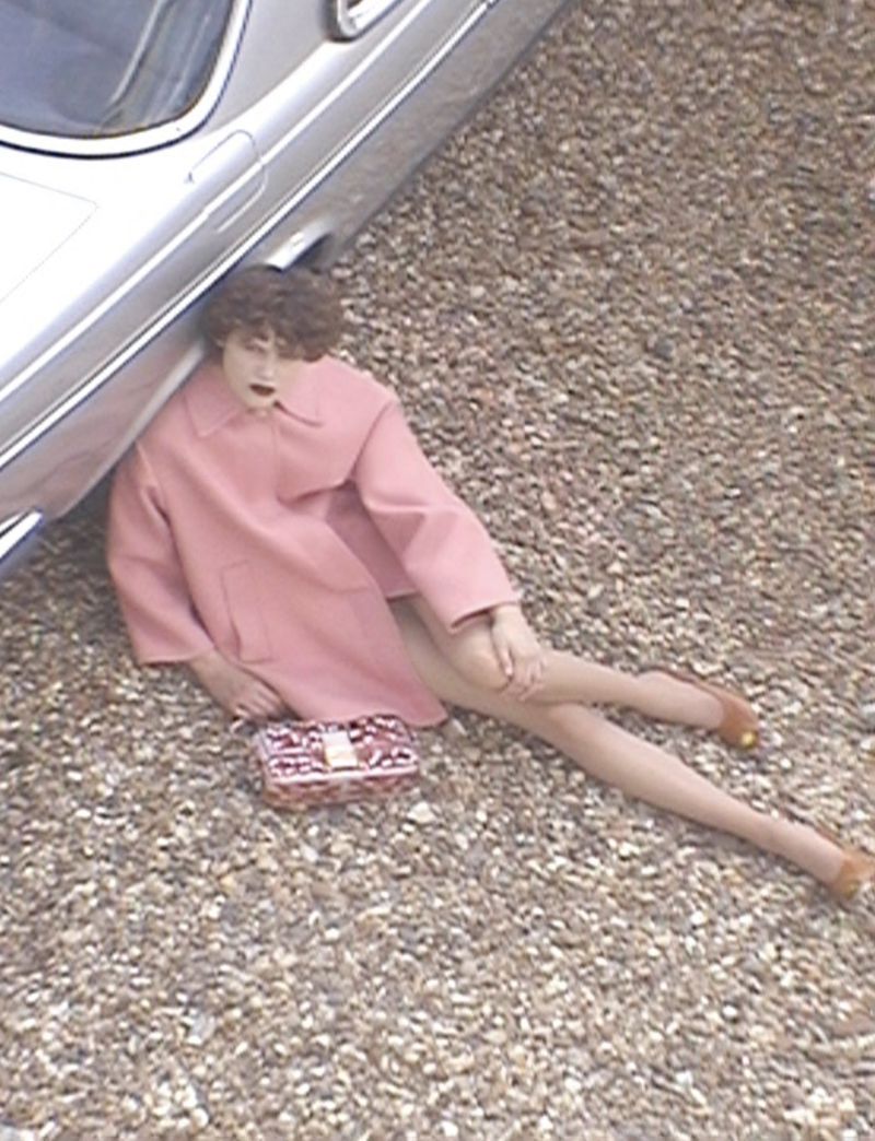 A person in a pink coat lies on pebbles next to a silver car, holding a pink and white bag, captured in a high-angle fashion shot.