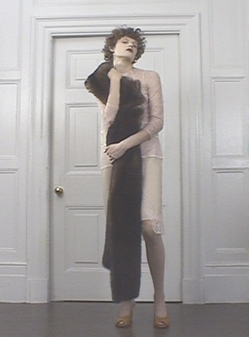 Woman in a light pink slip dress poses holding a dark brown faux fur stole in an indoor setting with a white door behind.