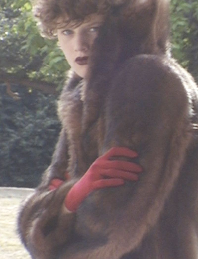 Woman wearing a large fur coat and red gloves standing outdoors with a background of trees and foliage in a fashion editorial style.