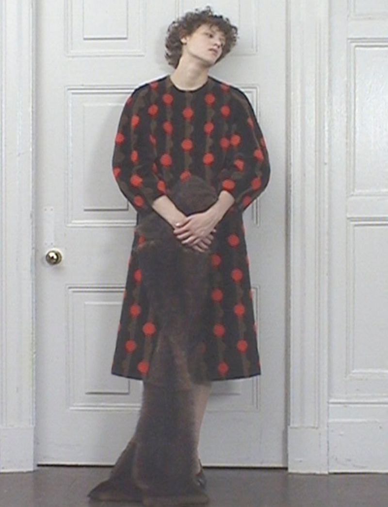 Adult female model wearing a brown dress with red polka dots posing in front of a white door inside a room with white walls.