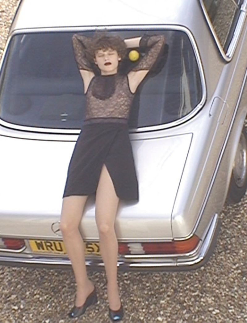 A woman in a lace top and skirt reclines against a vintage silver car outside, showcasing fashion and automotive elements.