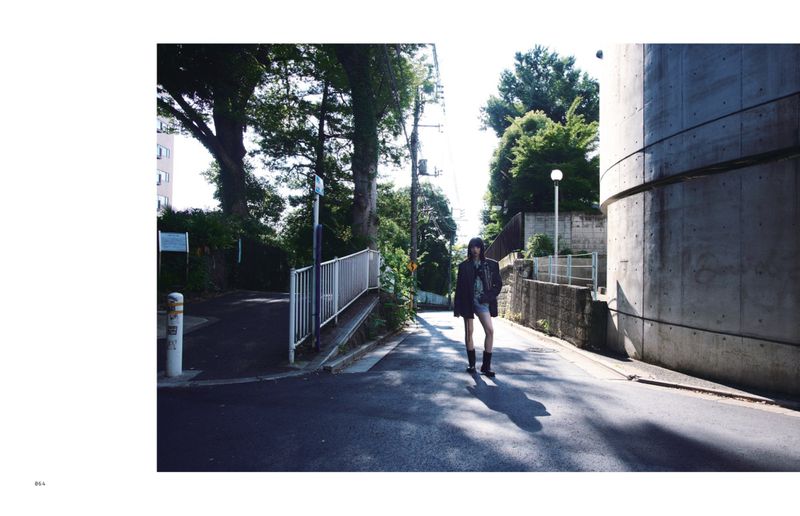 An adult female model walks on an asphalt street near a large concrete structure, trees, and foliage on a sunny day outdoors.
