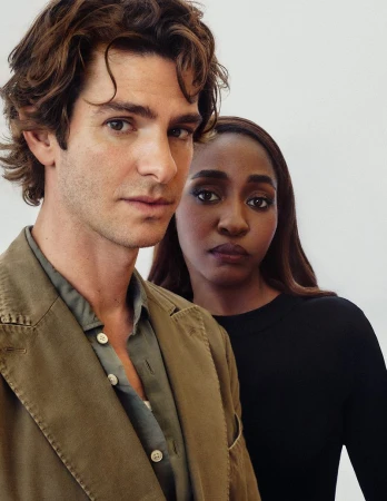 Headshot of Andrew Garfield, an adult male with brown wavy hair, standing next to a dark-skinned woman, shot in front of a plain white background.
