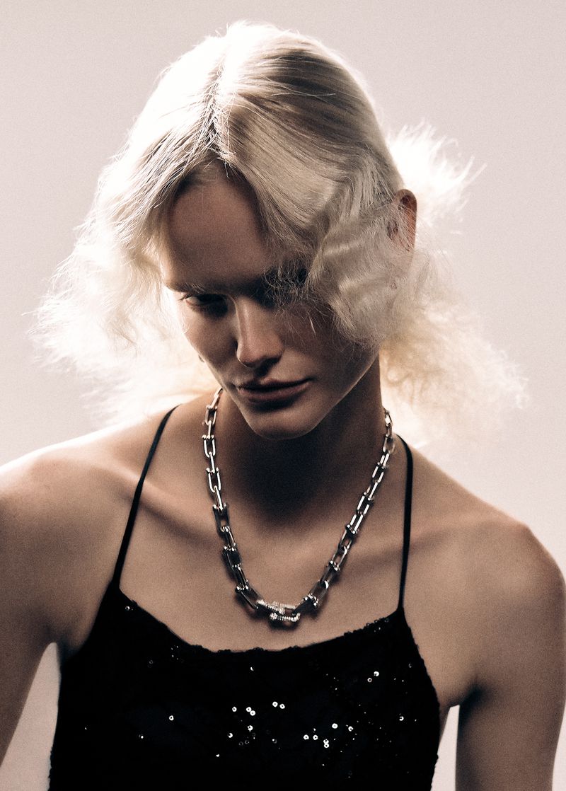 Close-up studio shot of a blond adult female fashion model wearing a silver necklace and spaghetti strap top, gazing downward.