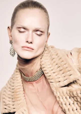 Woman with closed eyes poses in a beige fur coat with a gold necklace and earrings in a beauty fashion portrait in a studio.