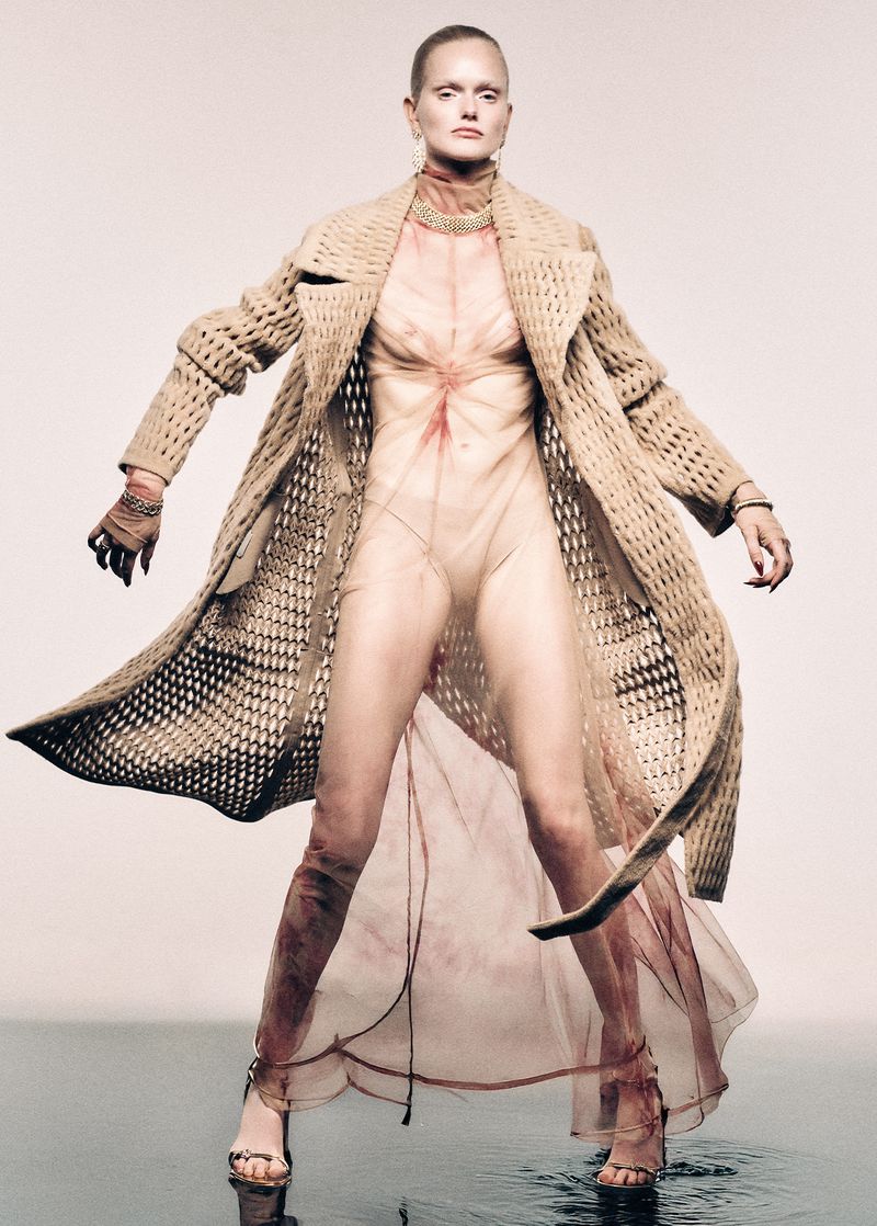 A slender female fashion model stands in a beige coat and sheer dress, striking a pose in a light, reflective studio environment with neutral tones.