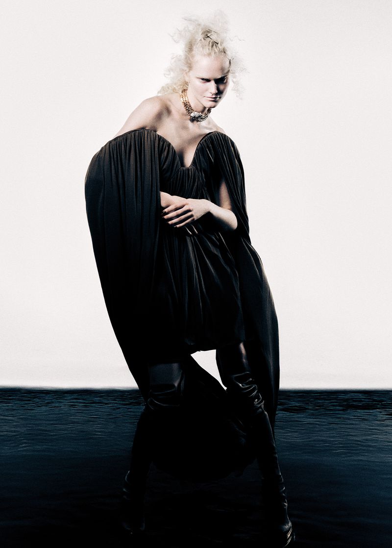 A woman in a black dress and high black boots poses against a white background with a strip of black water-like surface at the bottom.