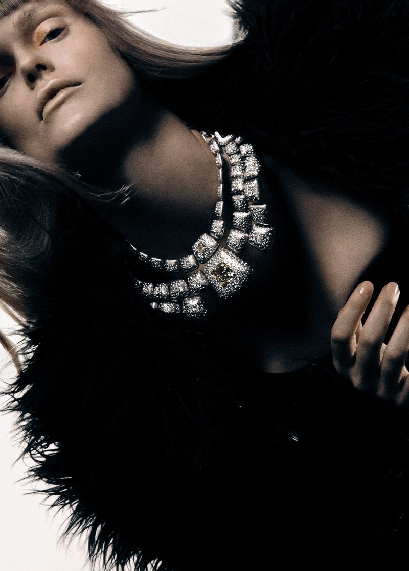 Close-up of a woman with fair skin and blonde hair wearing a large diamond necklace and a black feathered coat against a white backdrop.