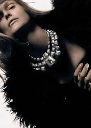 Close-up of a woman with fair skin and blonde hair wearing a large diamond necklace and a black feathered coat against a white backdrop.