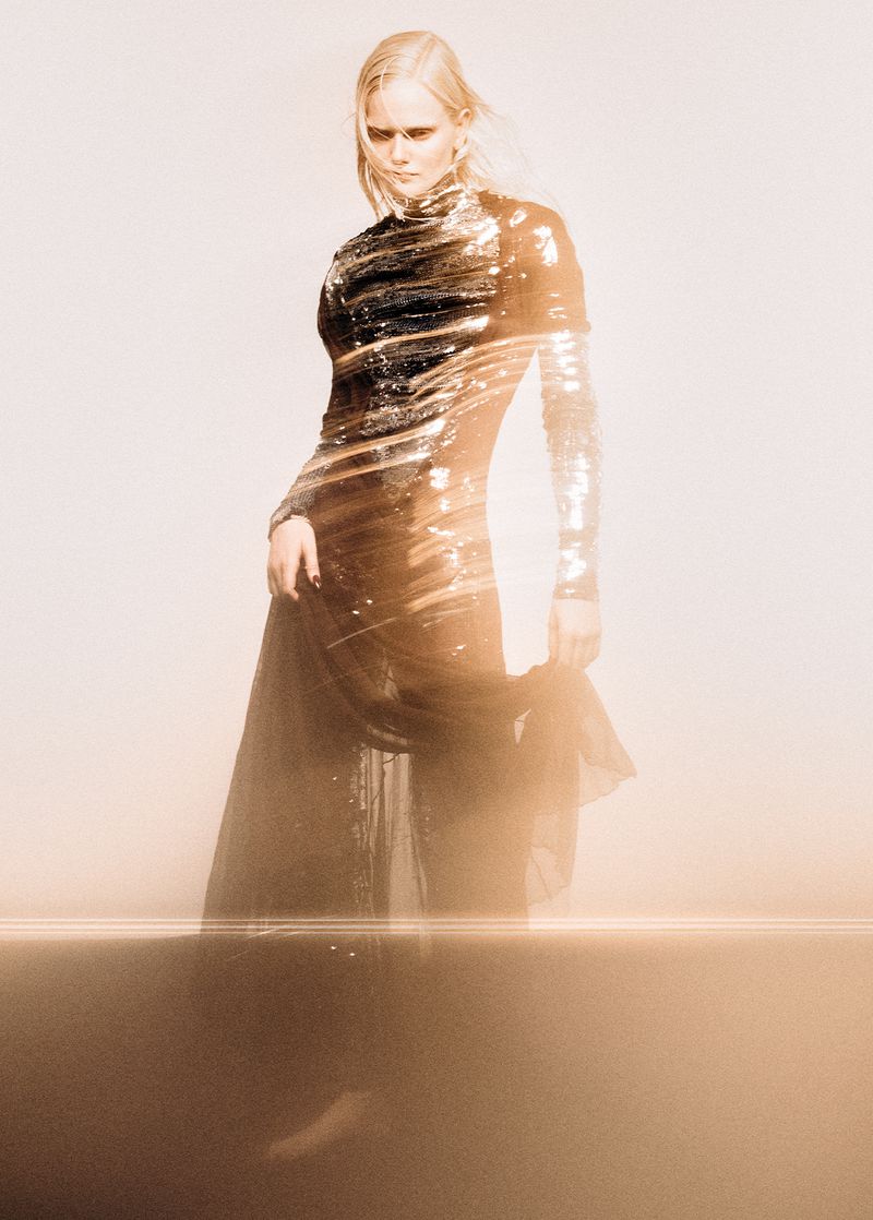 A full-length shot captures a fair-skinned adult female in a long, shimmering dress with long sleeves, in a photo studio setting.