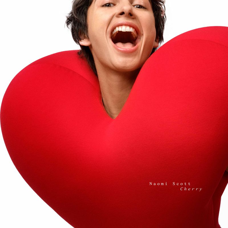 Person embracing a large red heart in a studio setting, smiling with mouth open. Close-up portrait against a clean, white backdrop.