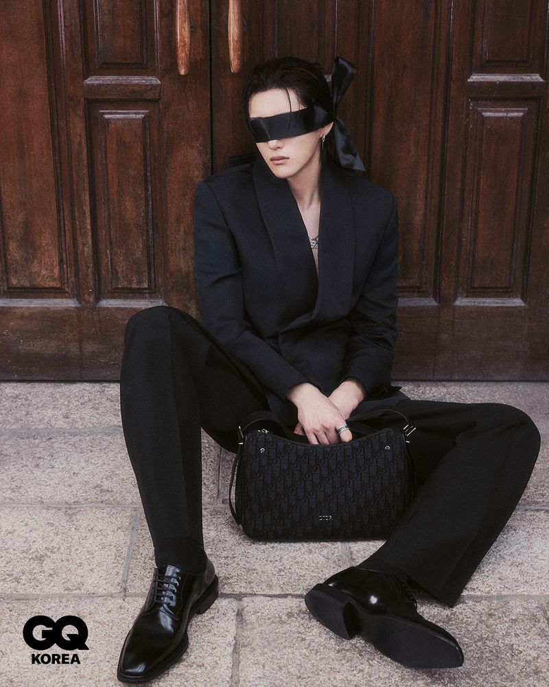 Portrait of a person in a black suit and blindfold sitting in front of a brown door with a black purse and shiny dress shoes.