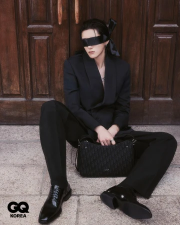 Portrait of a person in a black suit and blindfold sitting in front of a brown door with a black purse and shiny dress shoes.
