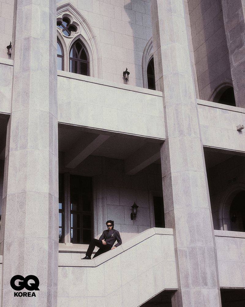 Fashion shot of a man in sunglasses and a jacket sitting on stairs, posing outdoors in front of a building with gothic architectural elements.