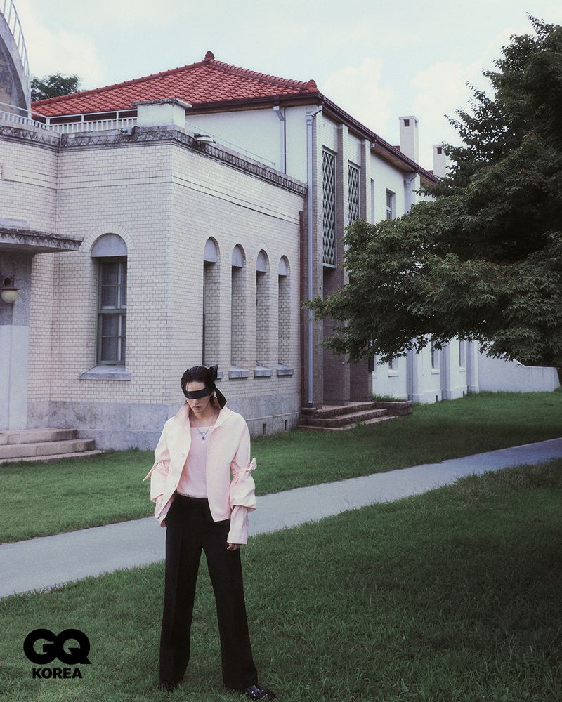A person wearing a blindfold, light pink jacket, and black pants stands on a grassy lawn in front of a light-colored building on a cloudy day.