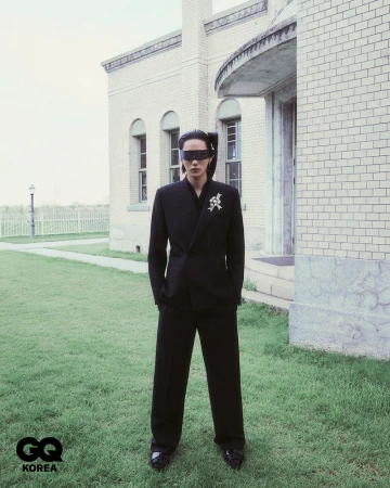 Adult male poses in a dark suit and geometric shades with a brick building and green lawn visible in the outdoor background.