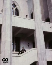 Model Hamin Yu wearing a dark suit and sunglasses seated on a stone staircase framed by massive pillars and arched windows.