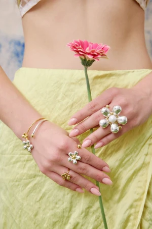 Close-up of a woman's hands adorned with flower-shaped jewelry, holding a pink gerbera daisy against a light green fabric background.