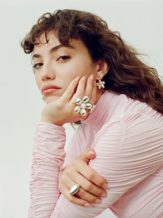 Brunette woman in pink turtleneck poses with flower jewelry including rings and earrings against a plain white backdrop in a studio.