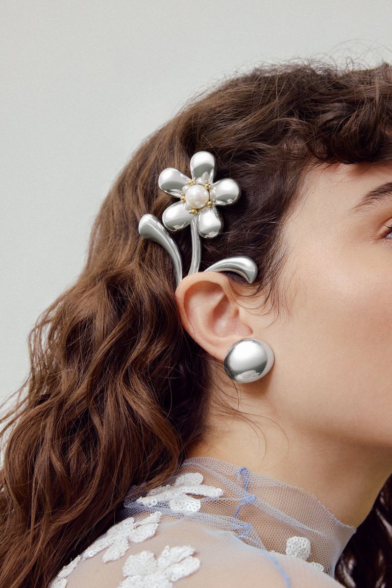 Close-up portrait of a woman wearing a silver metallic flower hair accessory and matching round earring against a neutral background.