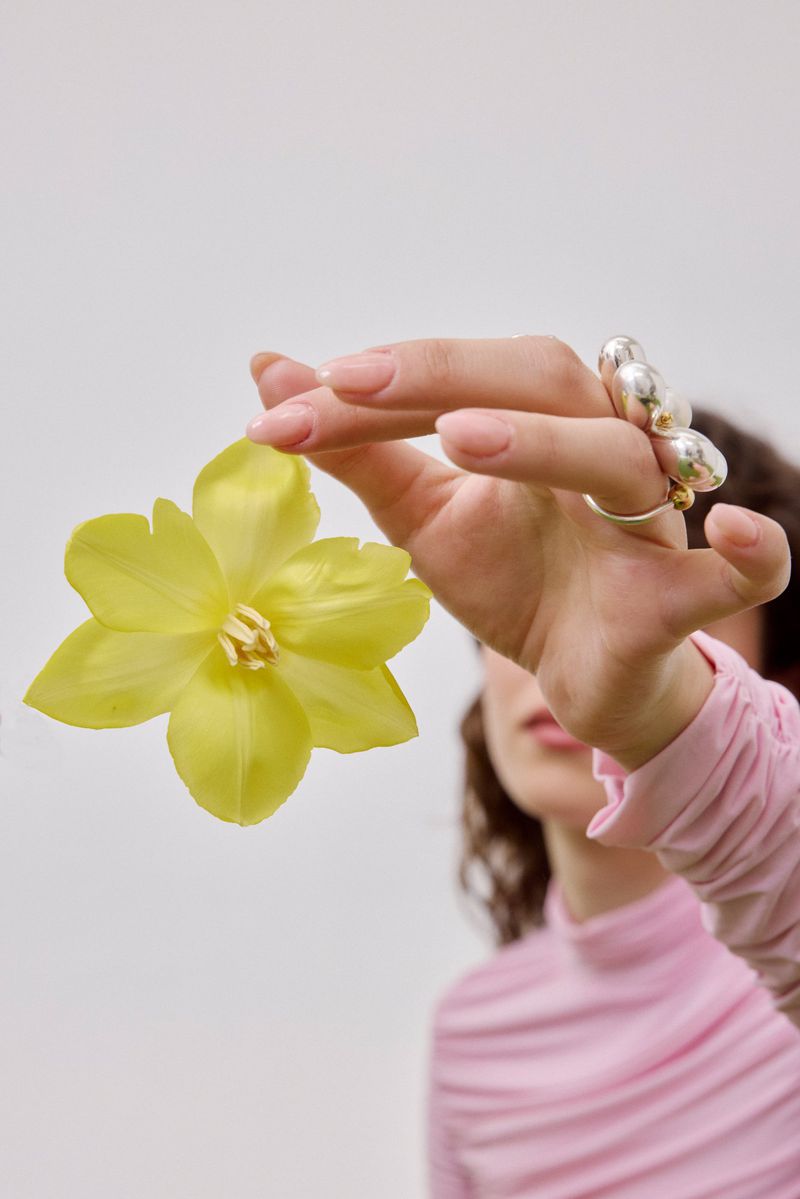 Woman's hand holding a large yellow flower, adorned with unique silver rings, against a soft pink top and a plain white background.