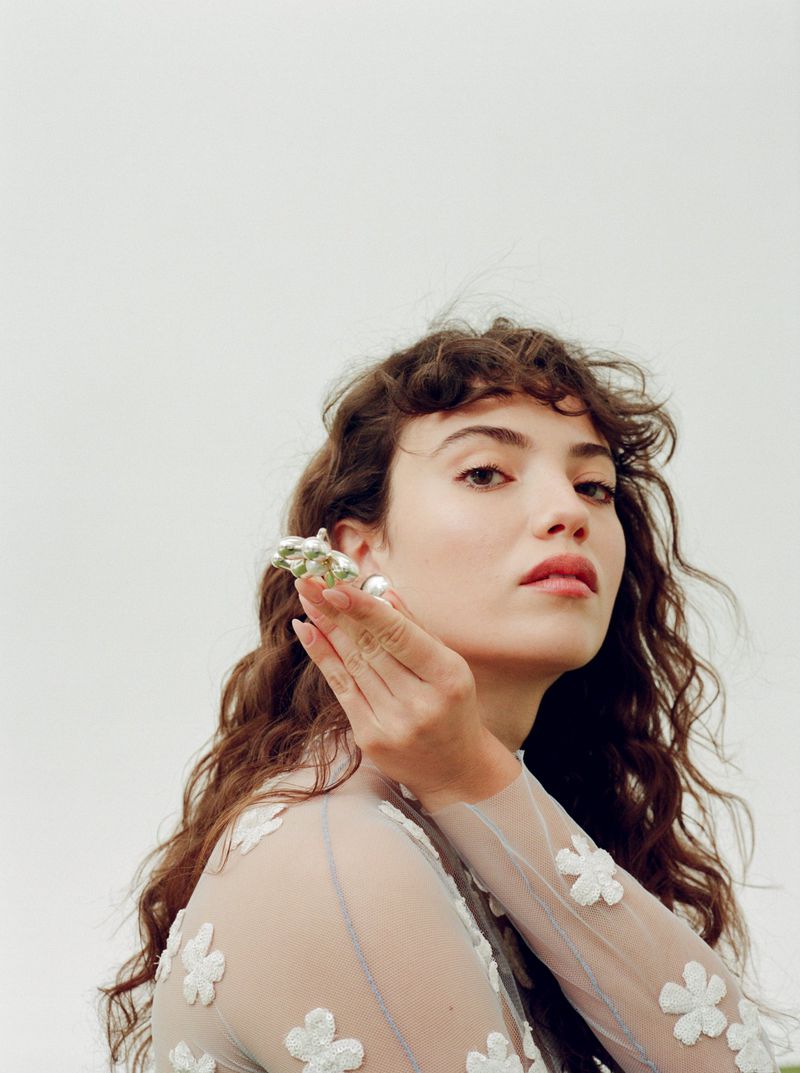 Woman with wavy brown hair and sheer flower-embellished dress, holding a seashell to her ear against a white backdrop with greenery.
