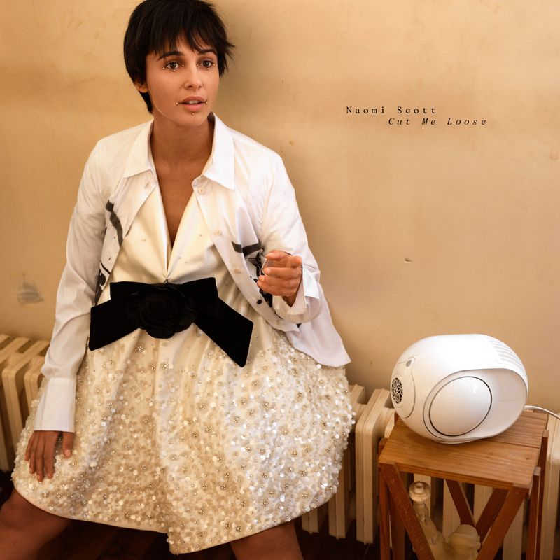 Naomi Scott poses in a white dress with a black bow, layered with an open white button-down shirt, next to a white speaker.