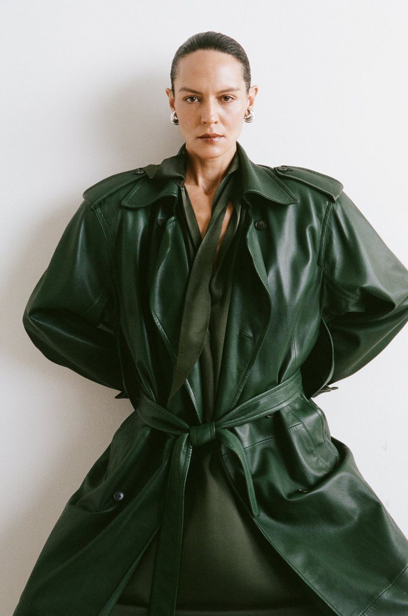 Woman in a green leather trench coat poses in front of a white wall; she wears black earrings and is styled in a low bun hairstyle.