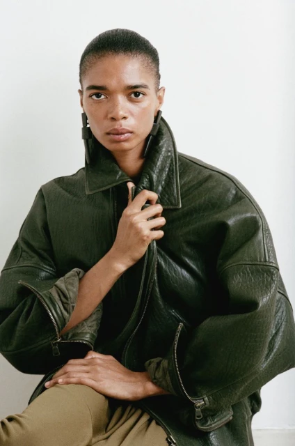 Adult female poses in a green leather bomber jacket against a neutral background, creating a modern and stylish fashion portrait.
