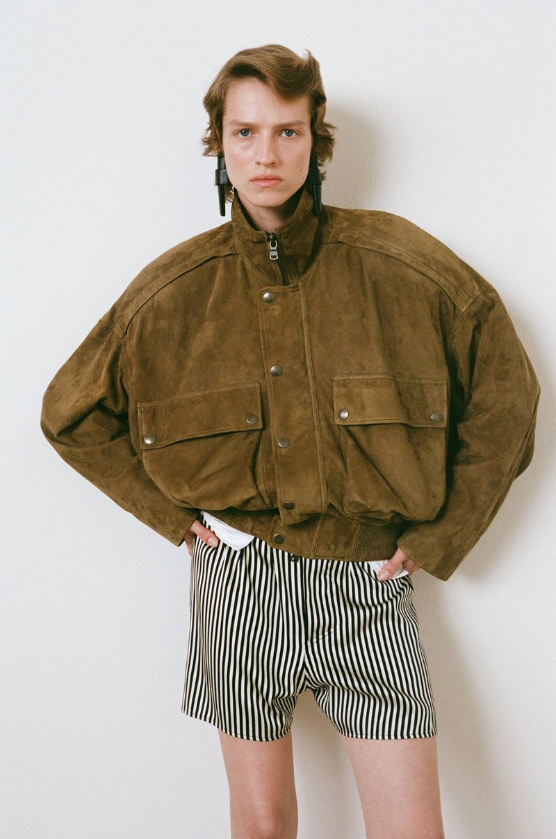 Woman in oversized brown suede jacket and striped shorts poses against a white background in a studio setting.