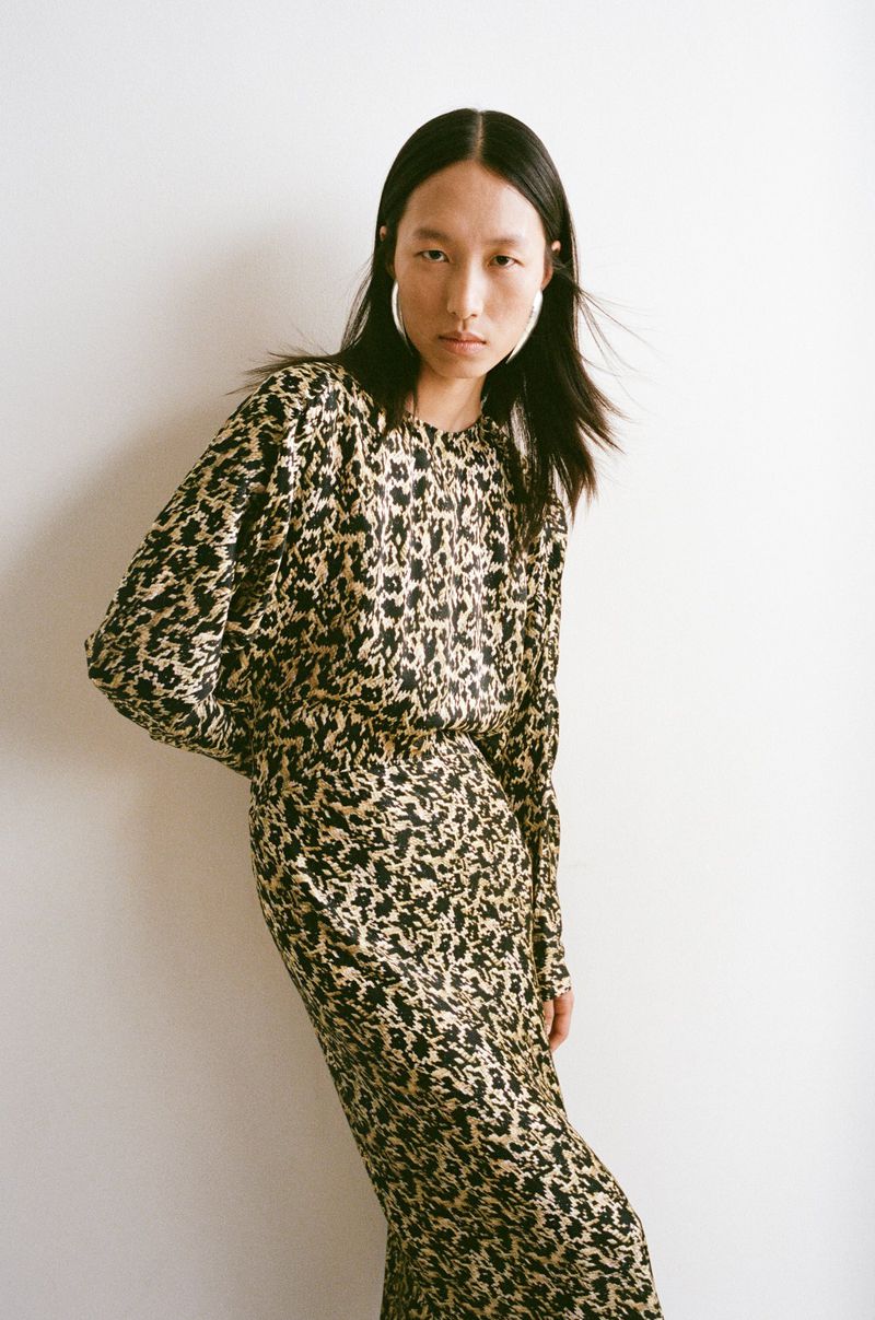 East Asian woman in a black and beige printed long-sleeve dress and large silver hoop earrings posing against a white background