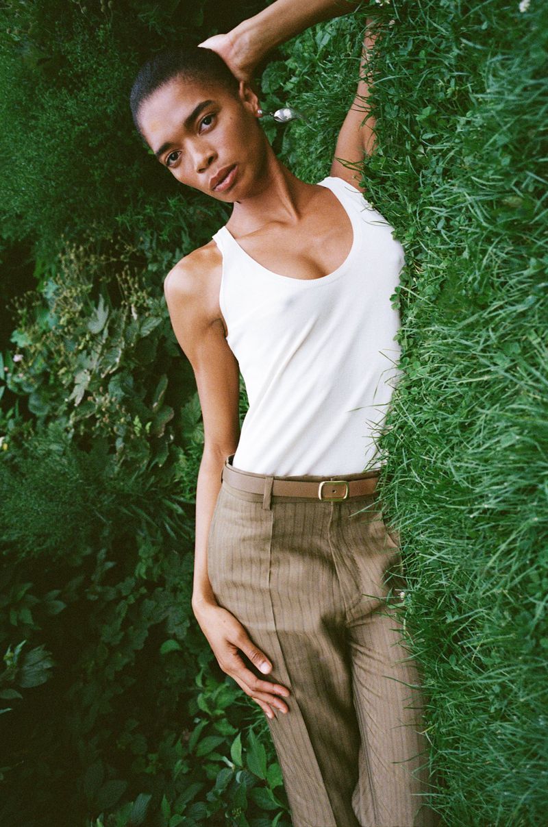 Model with buzzcut wearing a white tank top and tailored pinstripe trousers lying back against lush green foliage.