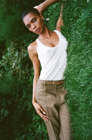 Model with buzzcut wearing a white tank top and tailored pinstripe trousers lying back against lush green foliage.