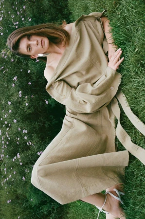 Model reclining on a grassy lawn wearing a draped beige dress and white strappy heels for the Petar Petrov lookbook.