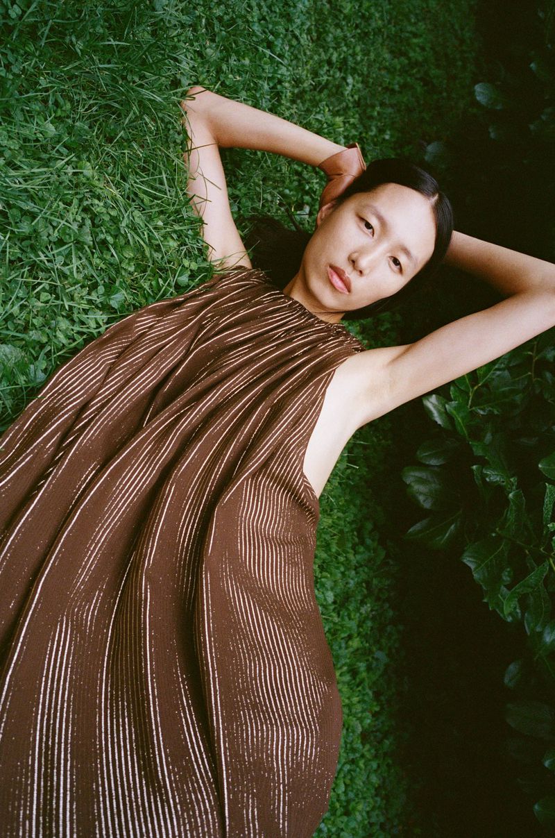 Young Asian woman lying on green grass, eyes open, wearing flowing brown dress, arms raised behind her head in outdoor setting.