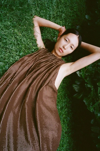 Young Asian woman lying on green grass, eyes open, wearing flowing brown dress, arms raised behind her head in outdoor setting.
