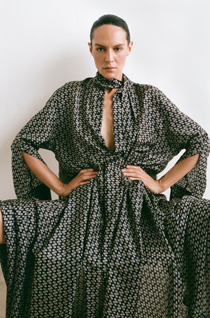 A woman wearing a black and white patterned dress is seated with her hands on her waist against a light background in a fashion shot.