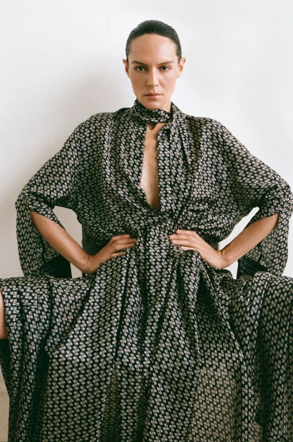 A woman wearing a black and white patterned dress is seated with her hands on her waist against a light background in a fashion shot.