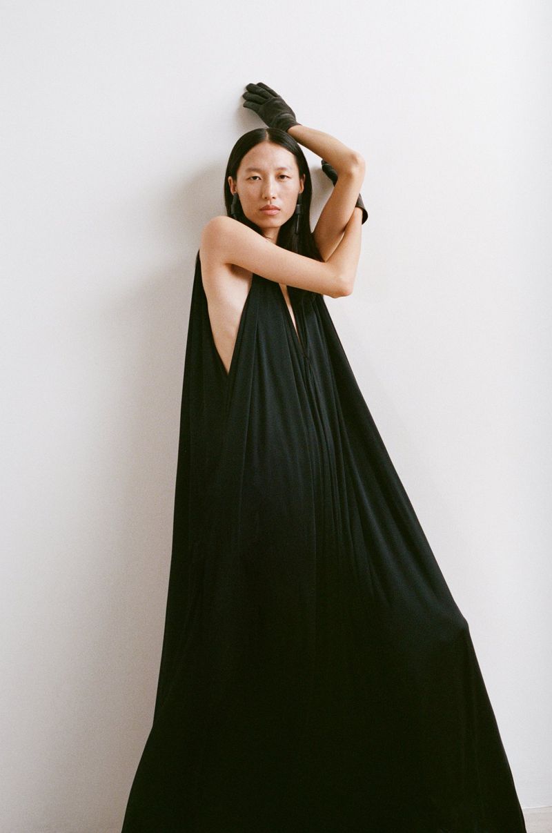 Model in a draped black halter maxi dress and black leather gloves posing against a white studio wall.
