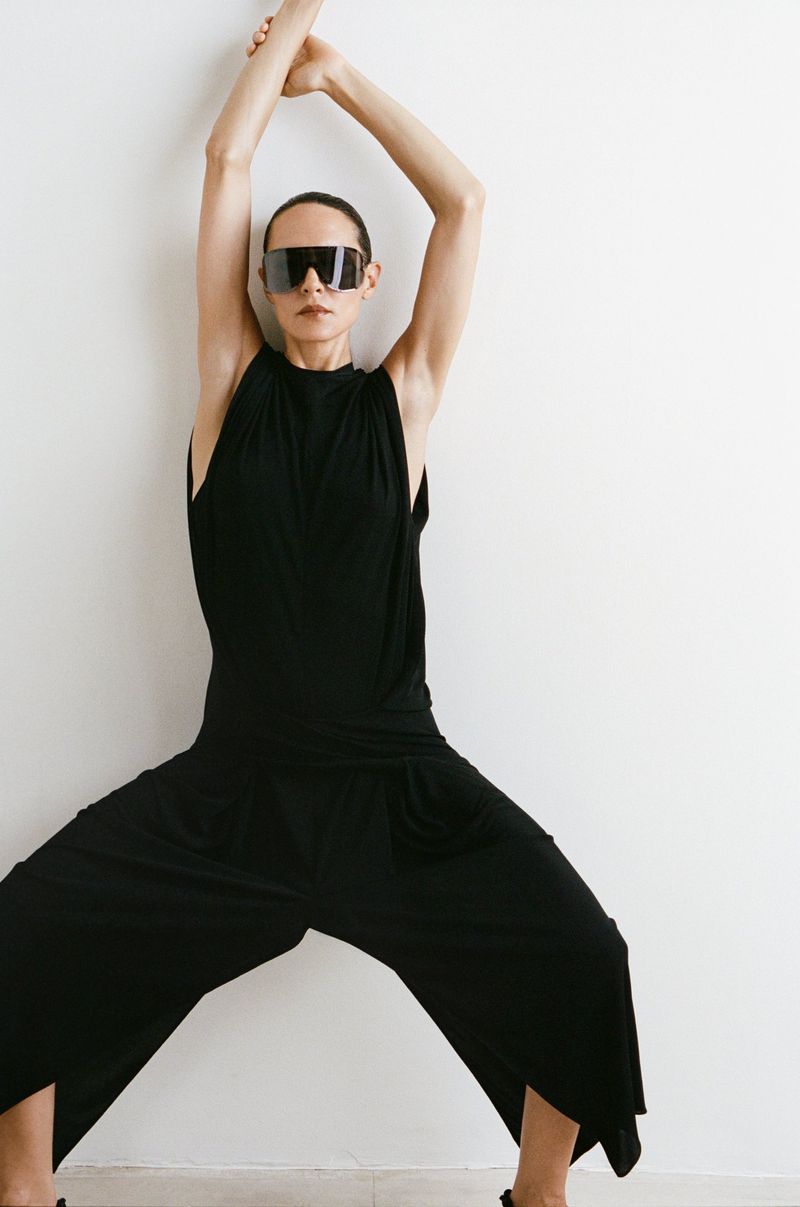 Woman in black jumpsuit and sunglasses poses against a white wall with arms raised overhead and legs spread wide, creating a bold silhouette.
