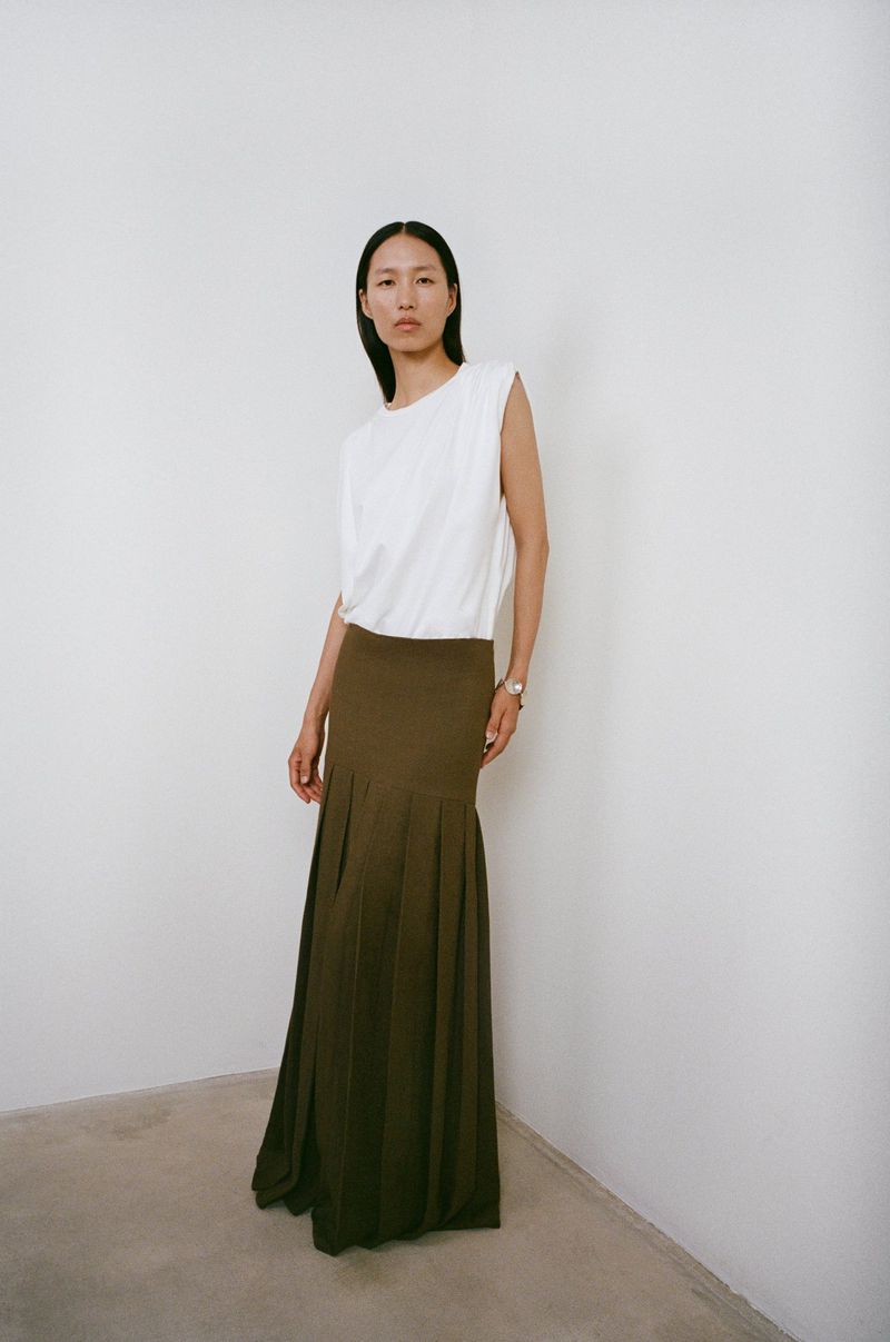 An Asian woman poses in a photo studio wearing a white top and flowing olive-green skirt, exuding modern and elegant style in a minimalist setting.