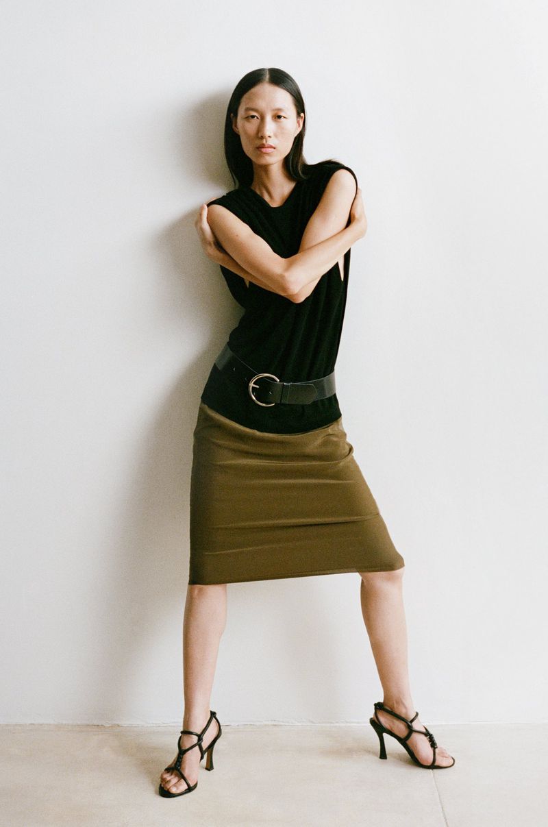Fashion shot of a young adult female model posing in a black top, tan skirt, and black strappy heels against a white wall.