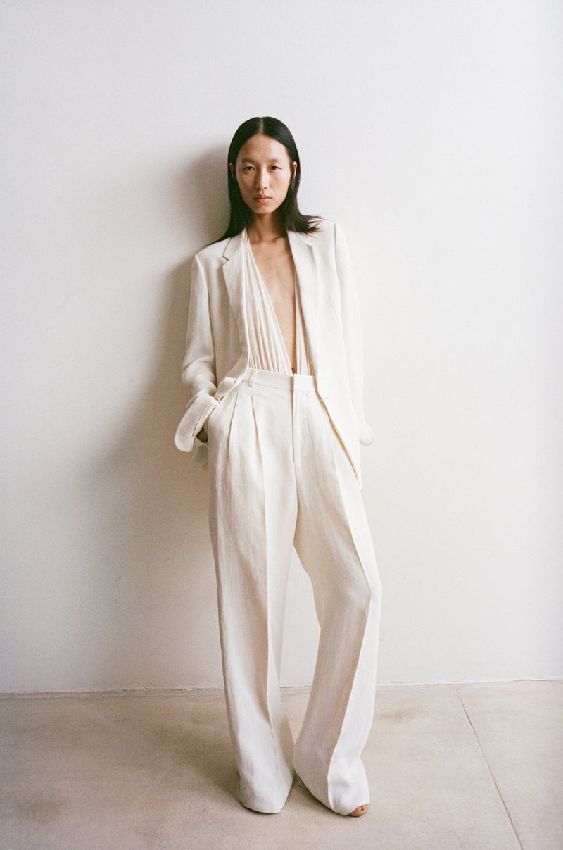 Fashion shot of a slender Asian woman in an unbuttoned white linen blazer and wide-legged pants, standing in front of a white wall.
