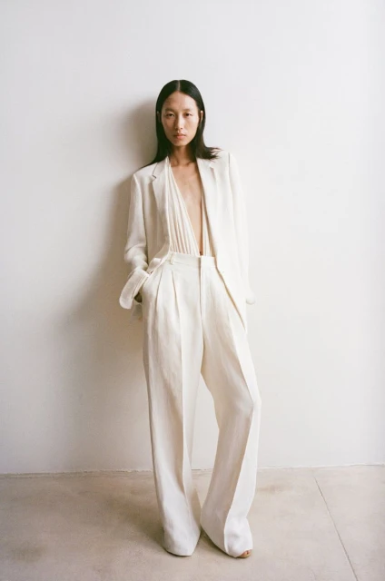 Fashion shot of a slender Asian woman in an unbuttoned white linen blazer and wide-legged pants, standing in front of a white wall.