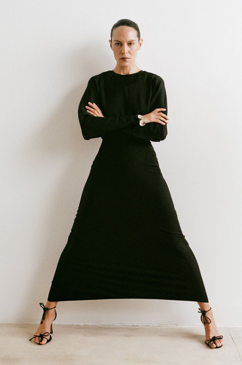 Elegant woman in a long black dress stands with crossed arms against a white wall, showcasing a minimalist fashion aesthetic.